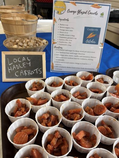 A food display features many small paper cups filled with cooked carrot pieces, a recipe sign, and a small chalkboard reading 'Local Hadley Carrots.'